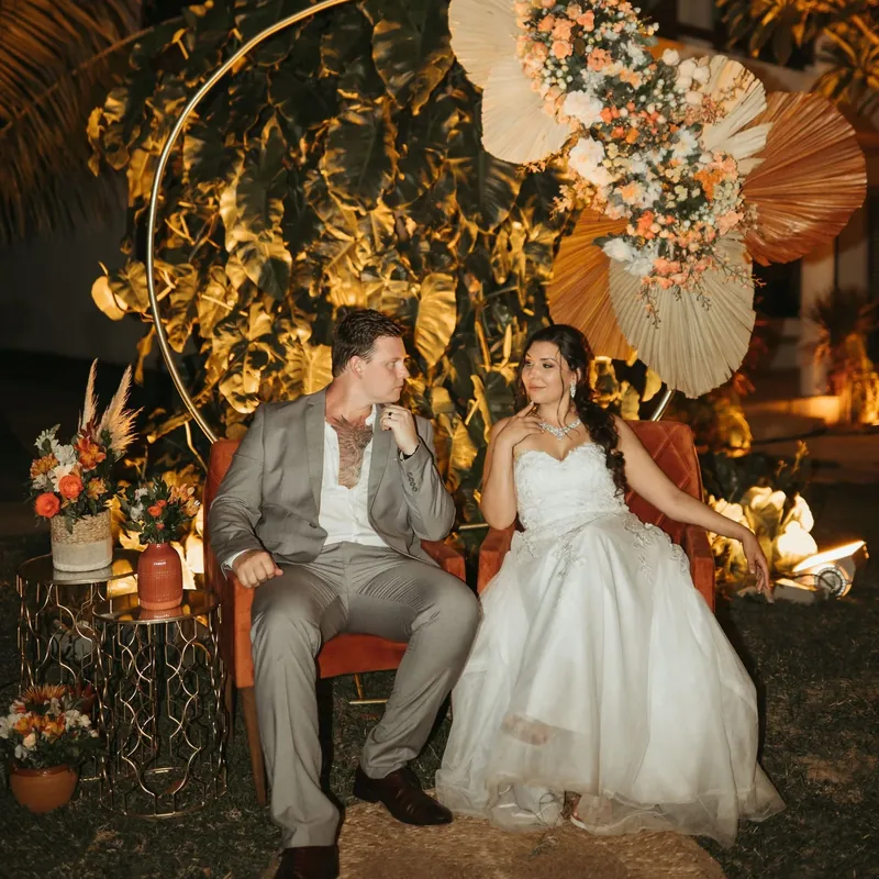 A just-married couple sitting together at their reception under palms and warm light.